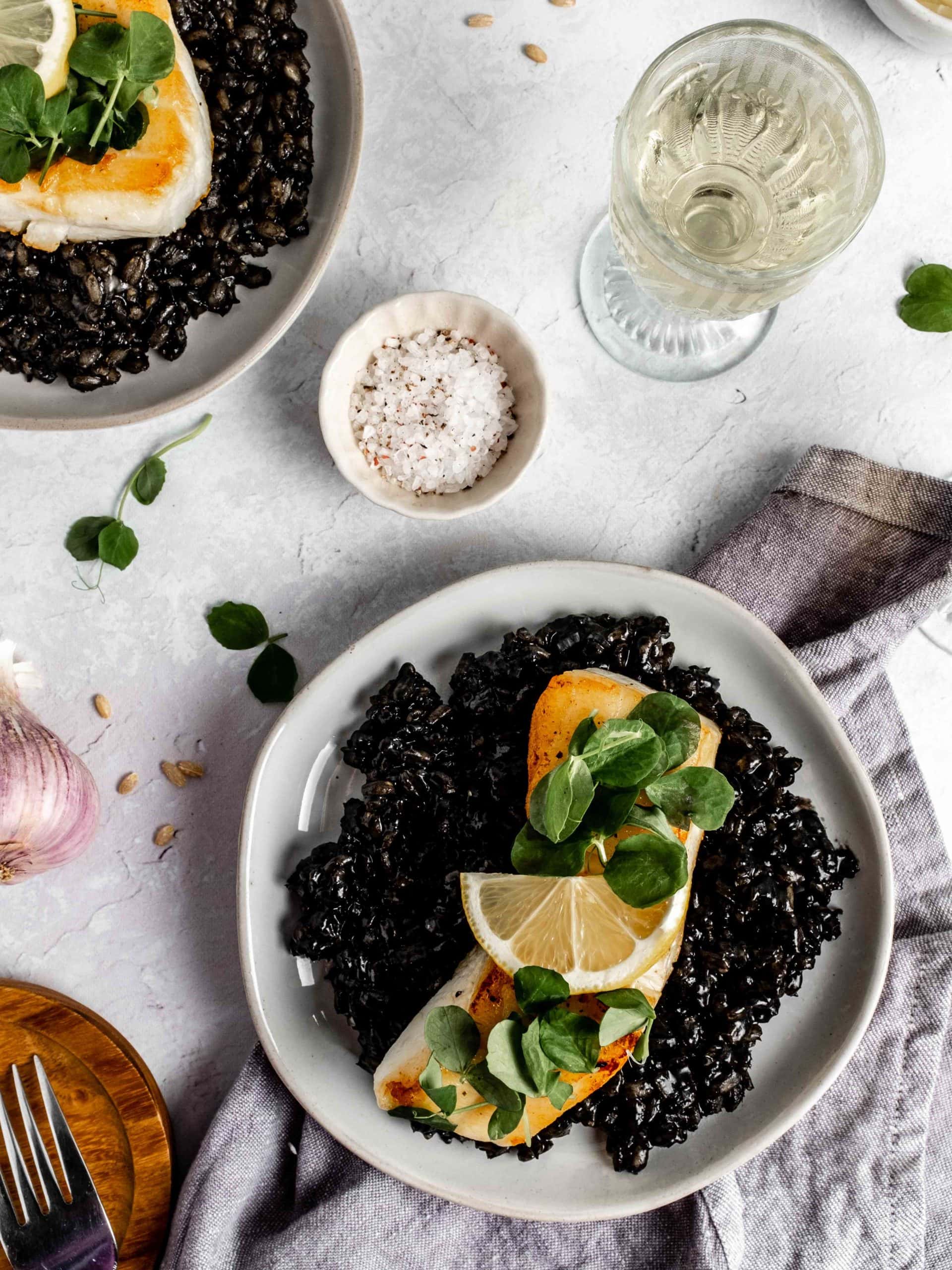 two plates of squid ink barley risotto topped with seared sea bass and lemon