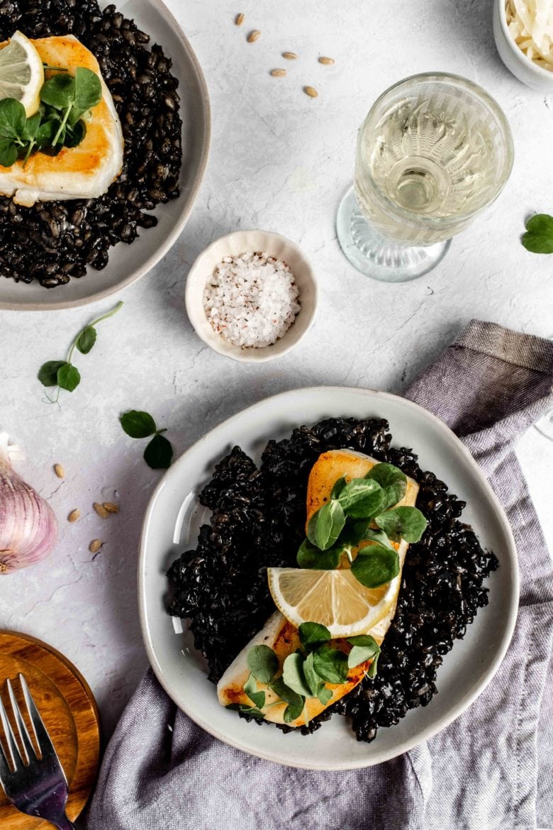 two plates of squid ink barley risotto topped with seared sea bass and lemon