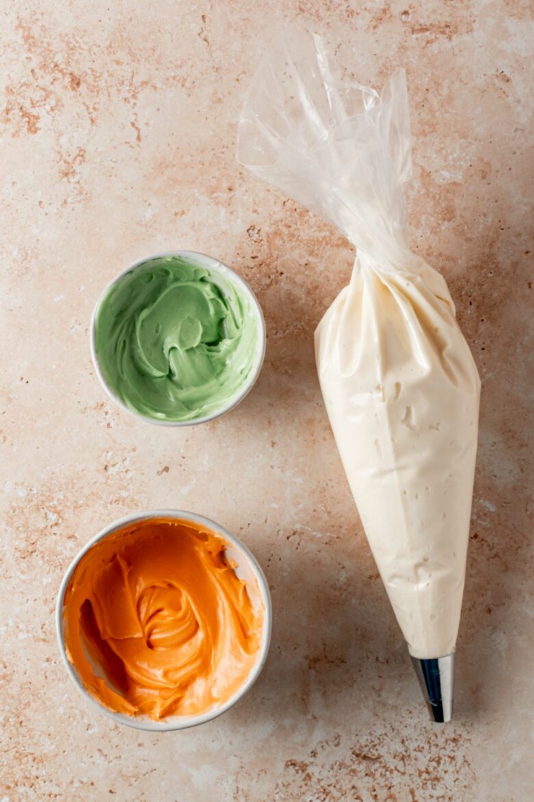 Piping bag with white frosting and two small bowls with orange and green frosting