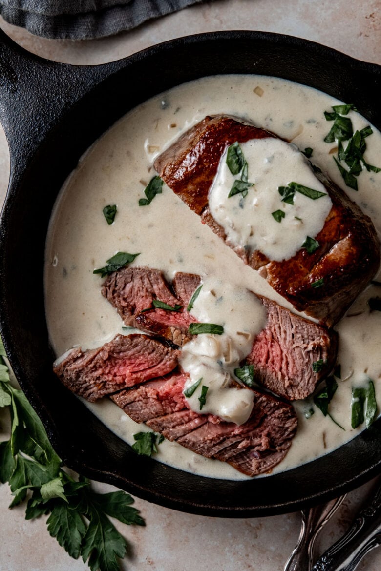 Filet mignon slices in a small cast iron skillet topped with creamy blue cheese sauce and garnished with fresh parsley.