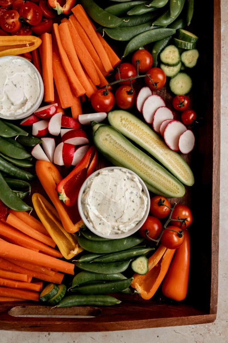 Assorted vegetables arranged on wooden serving tray with dips in small bowls.