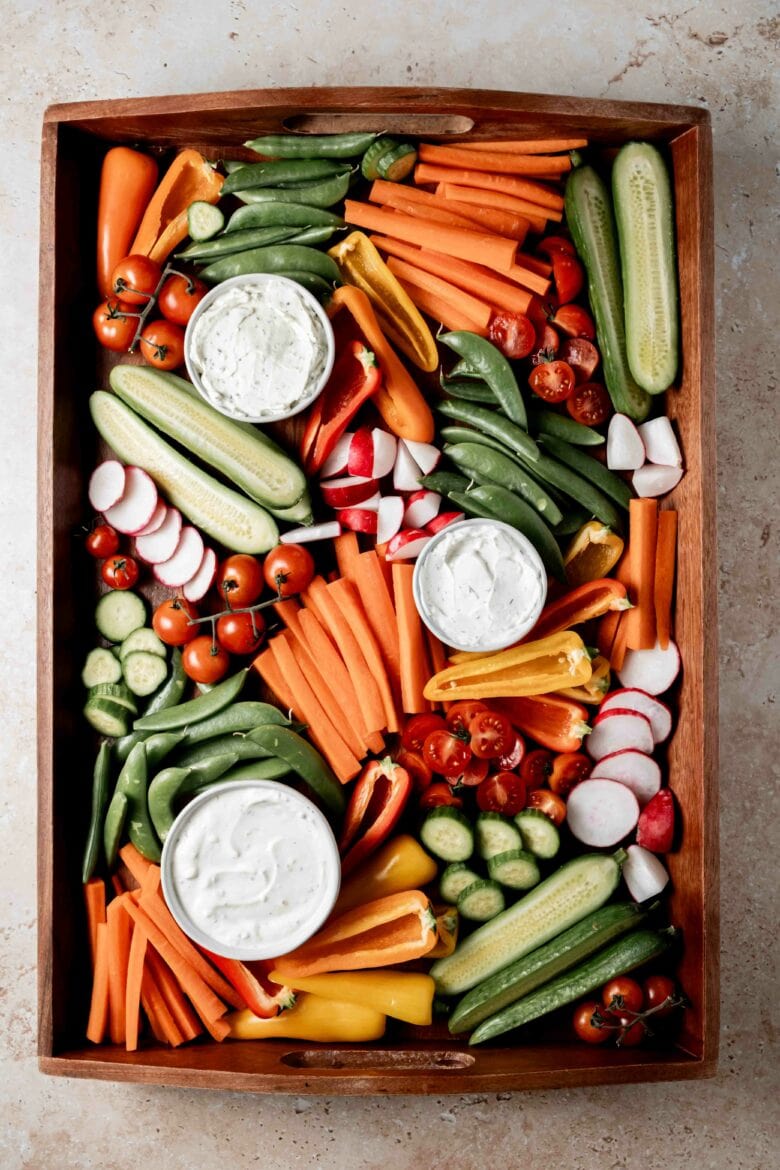 Completed platter with cucumbers, carrots, snap peas, radishes, tomatoes and bell peppers alongside three dips.