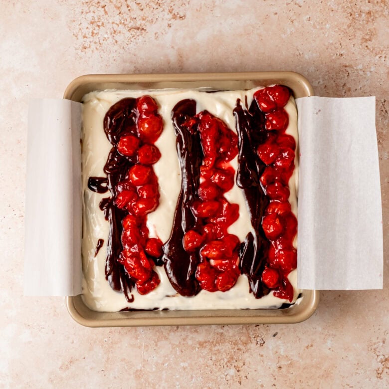 Brownies with lines of cheesecake, brownie and cherries before swirling.